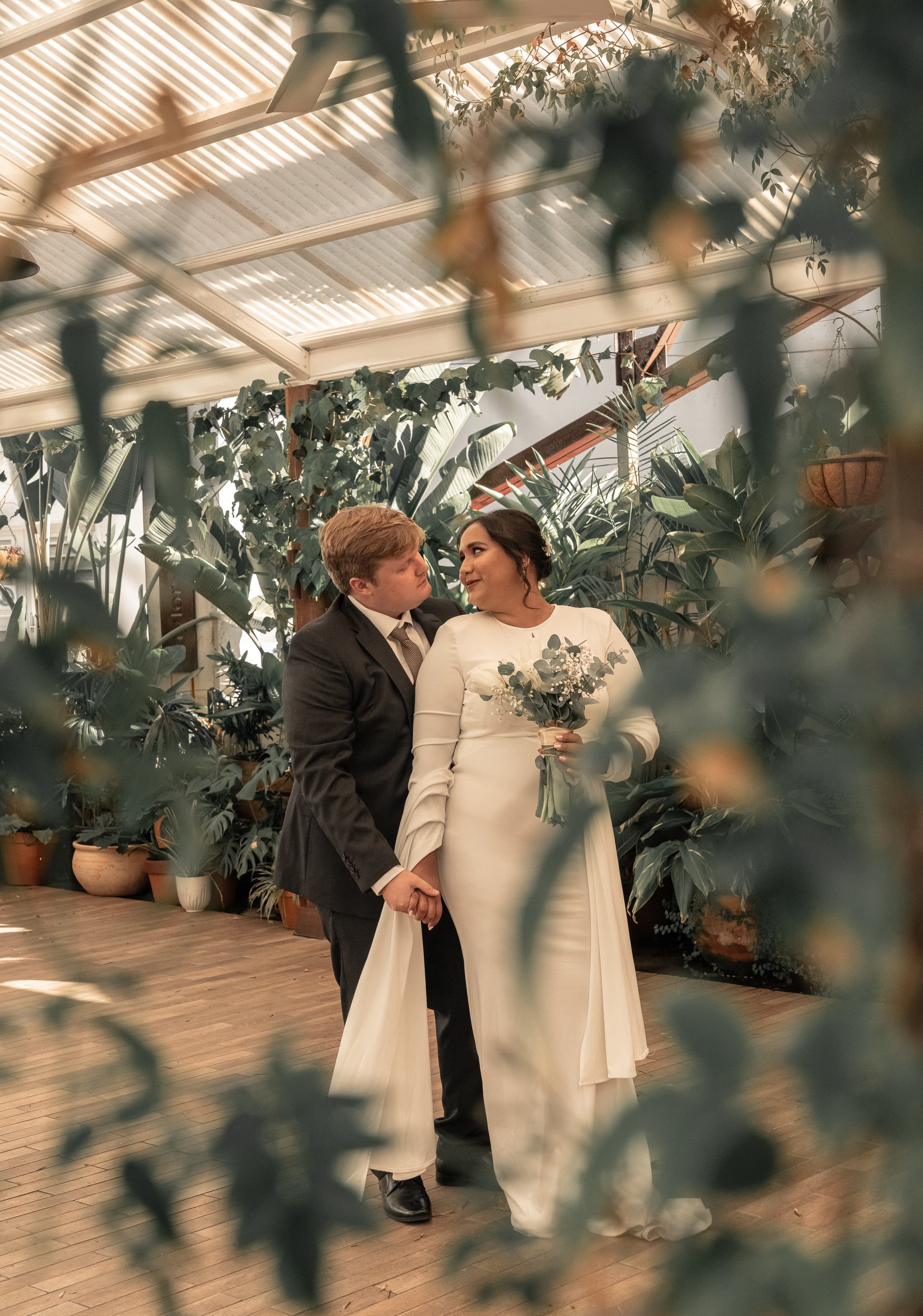 Bride and groom gaze lovingly at each other in a lush, sunlit greenhouse conservatory.