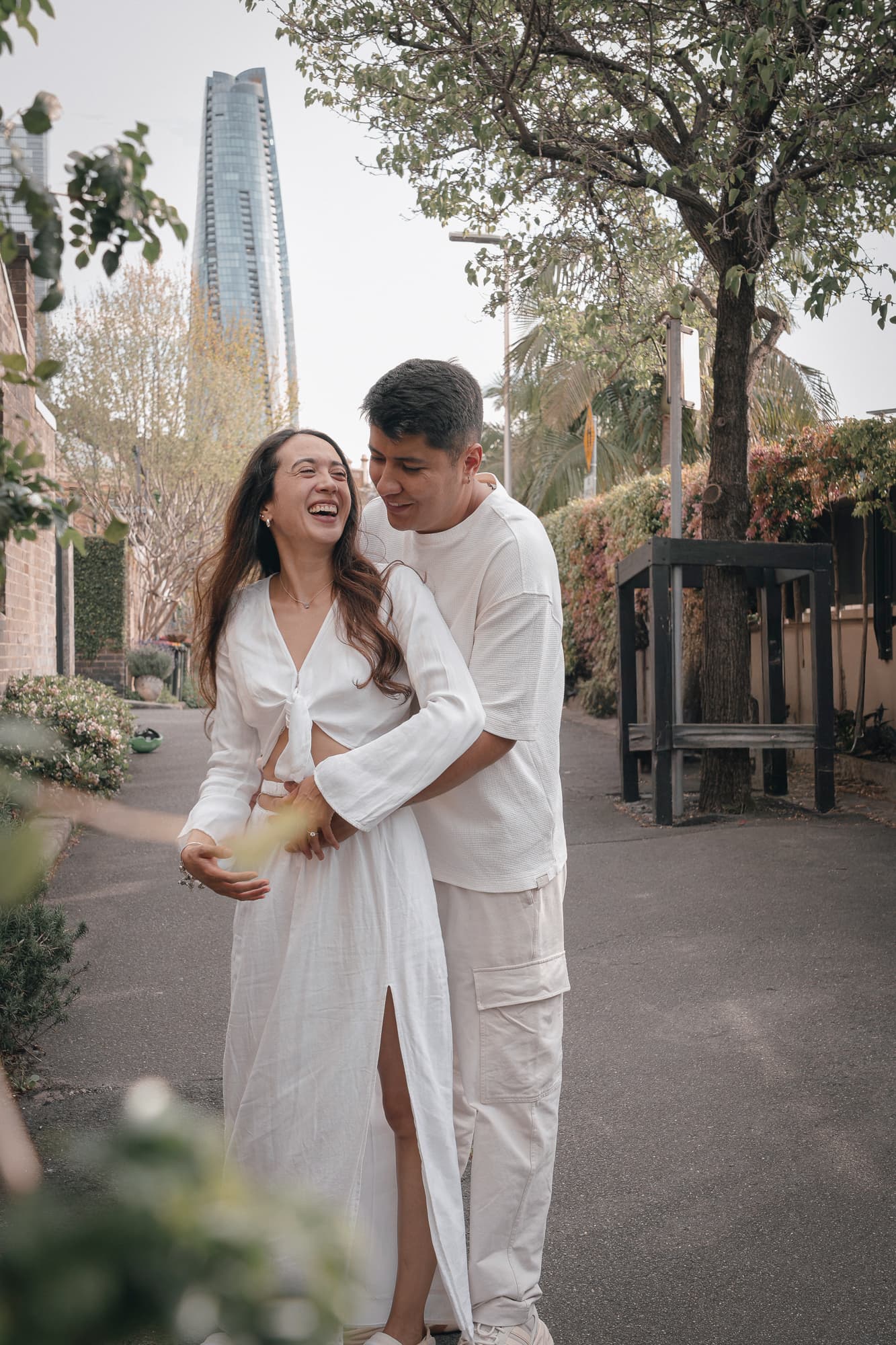 Laughing couple in white outfits embraces on a city street with a skyscraper behind.