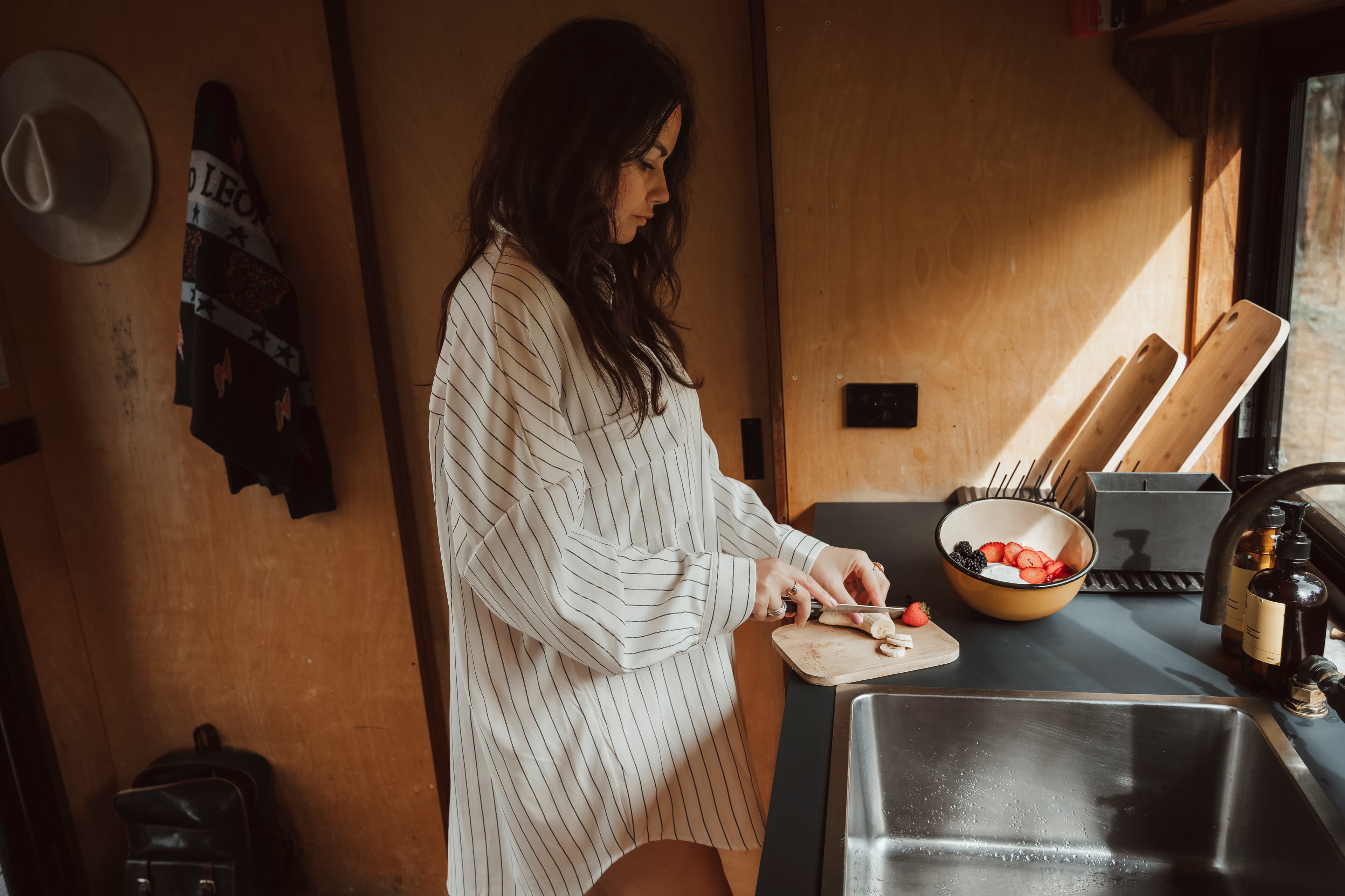 Woman in a pinstriped shirt slices a banana in a sunlit, rustic wooden kitchen.