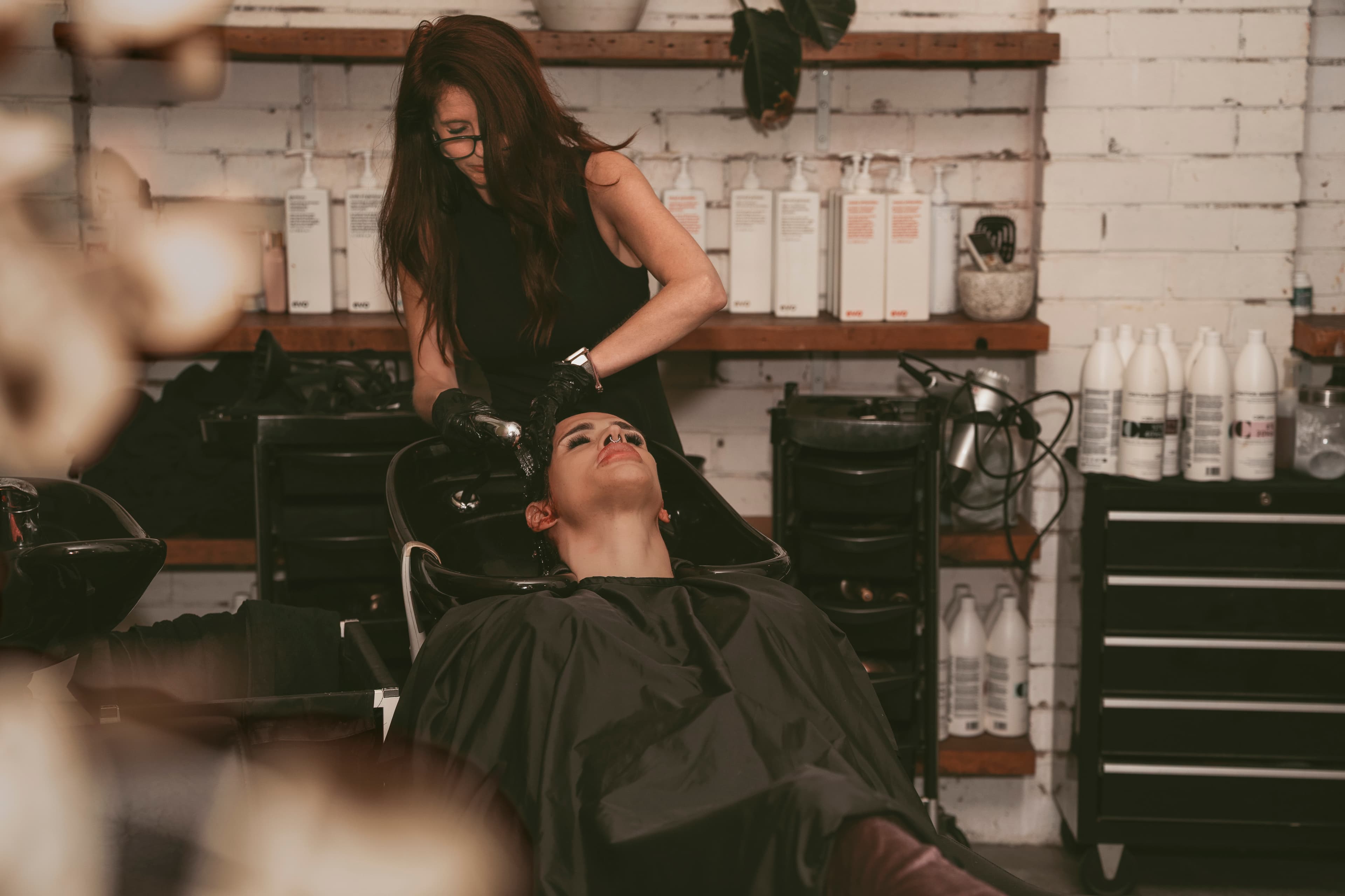 A hairstylist wearing black gloves washes a client's hair in a black salon sink.
