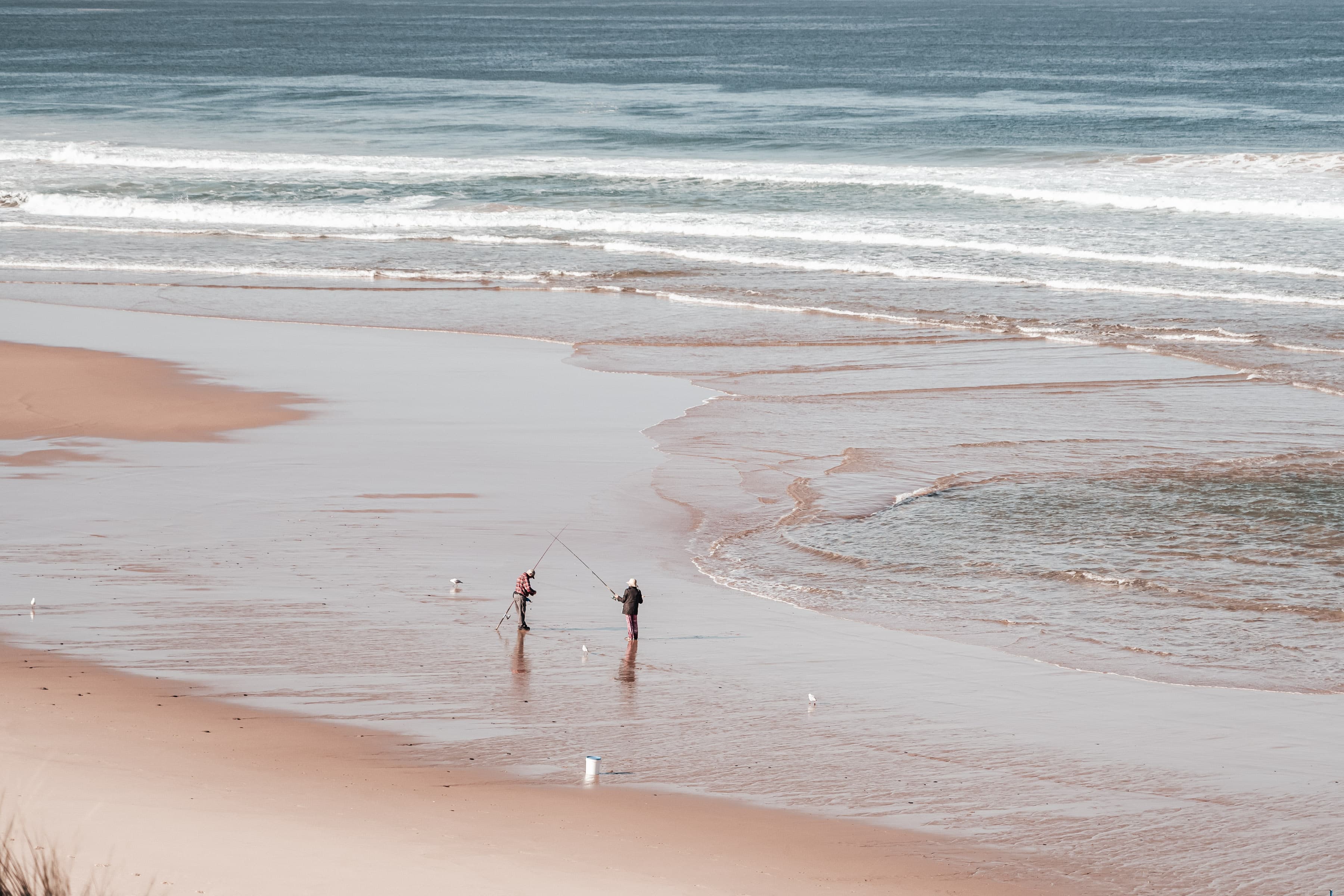 Two people surf fishing on a vast sandy beach with gentle waves rolling ashore.