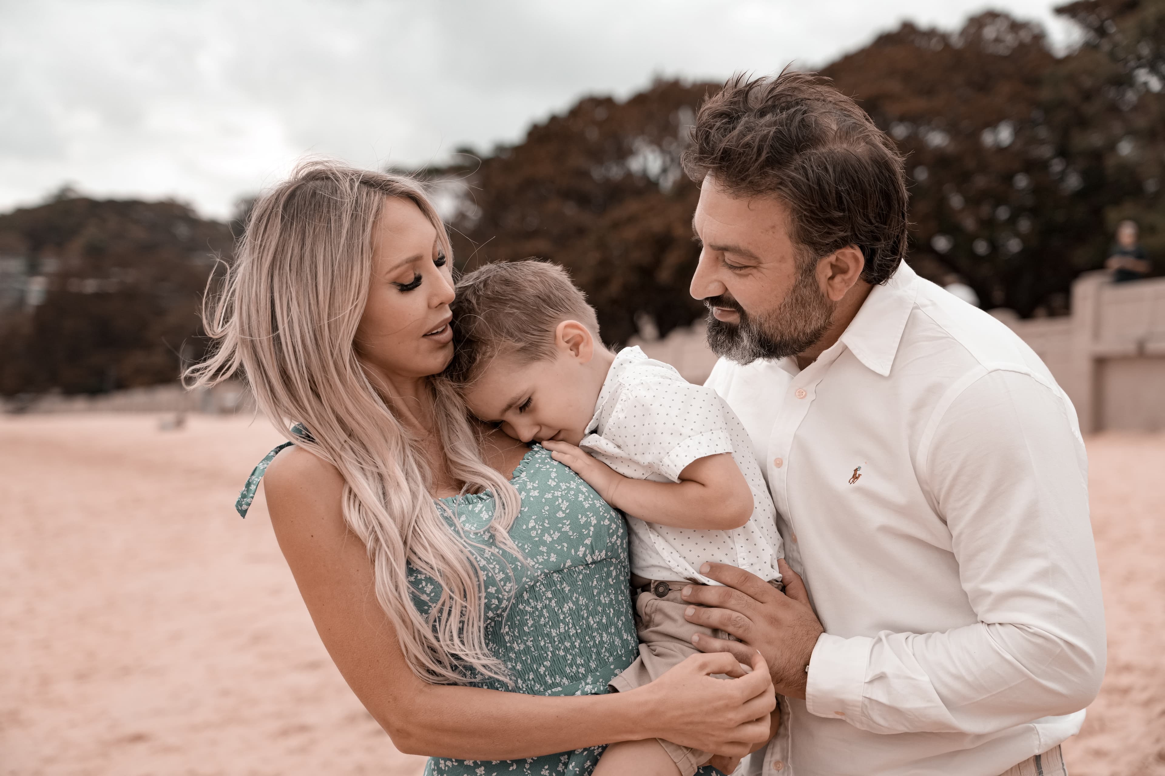 A blonde woman and bearded man hold their young son closely on a sandy beach.