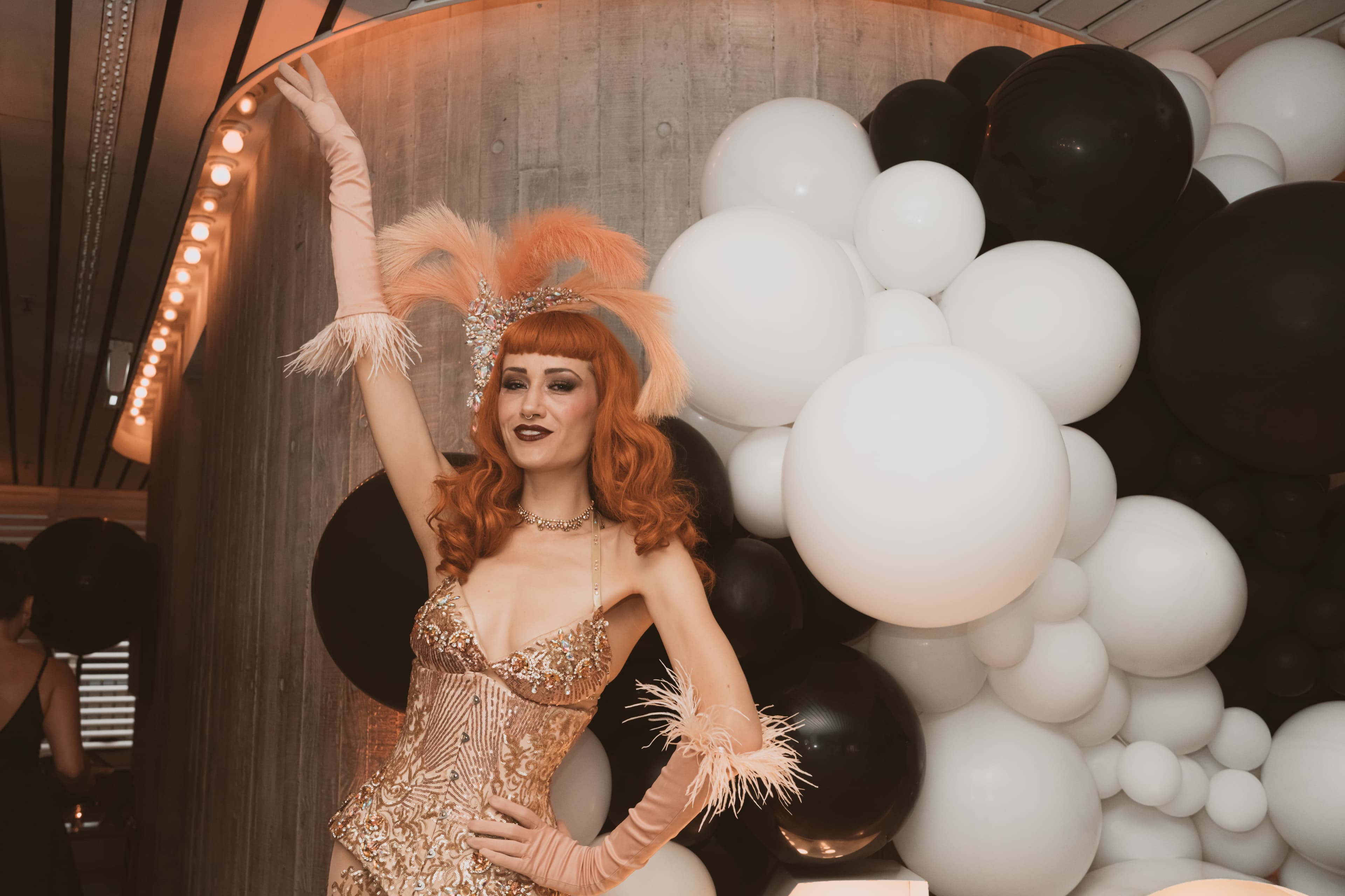 Red-haired showgirl in gold sequins posing against a backdrop of black and white balloons.