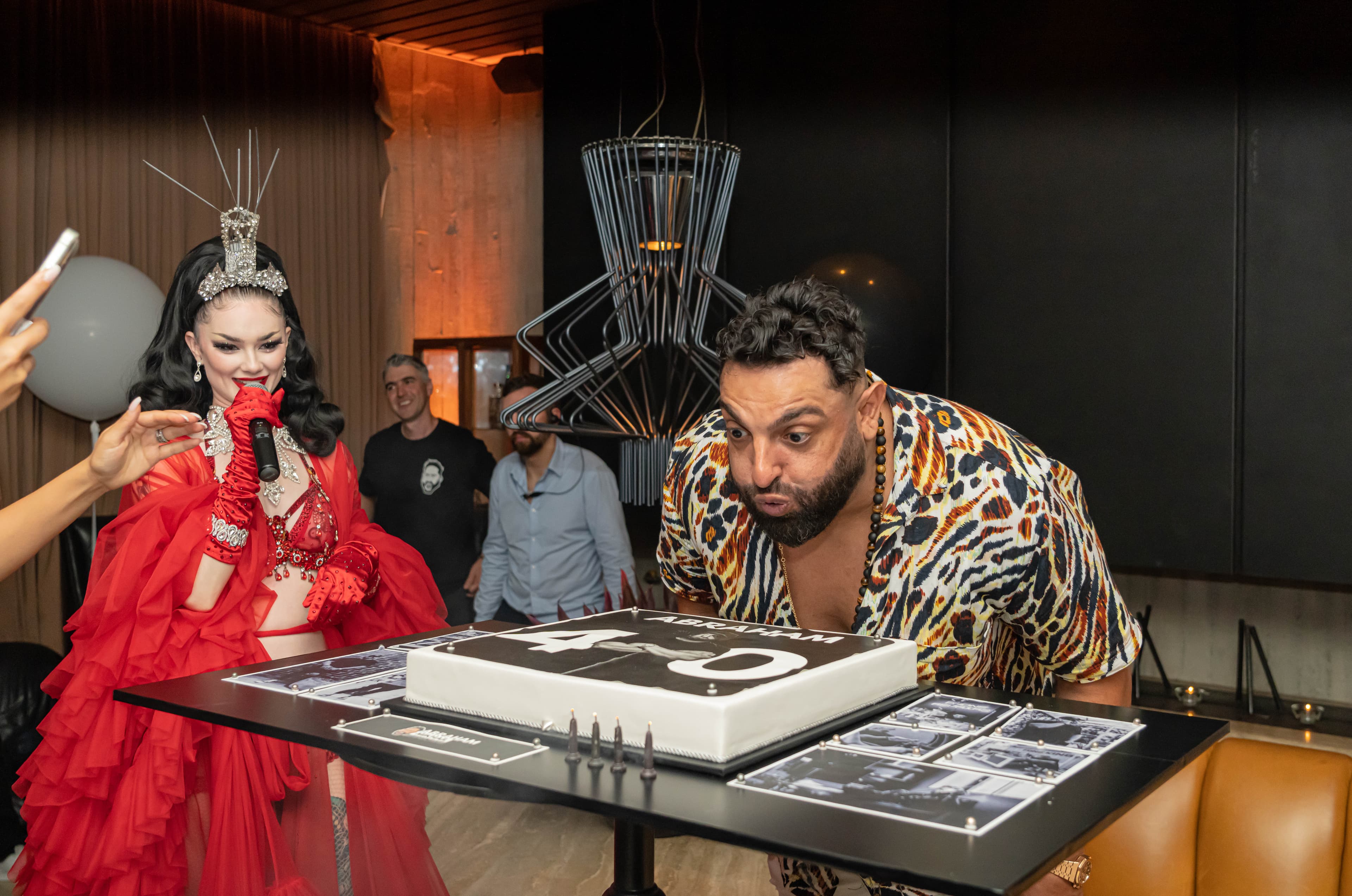 Man in leopard print shirt blows out candles on a 40th birthday cake beside a drag queen.