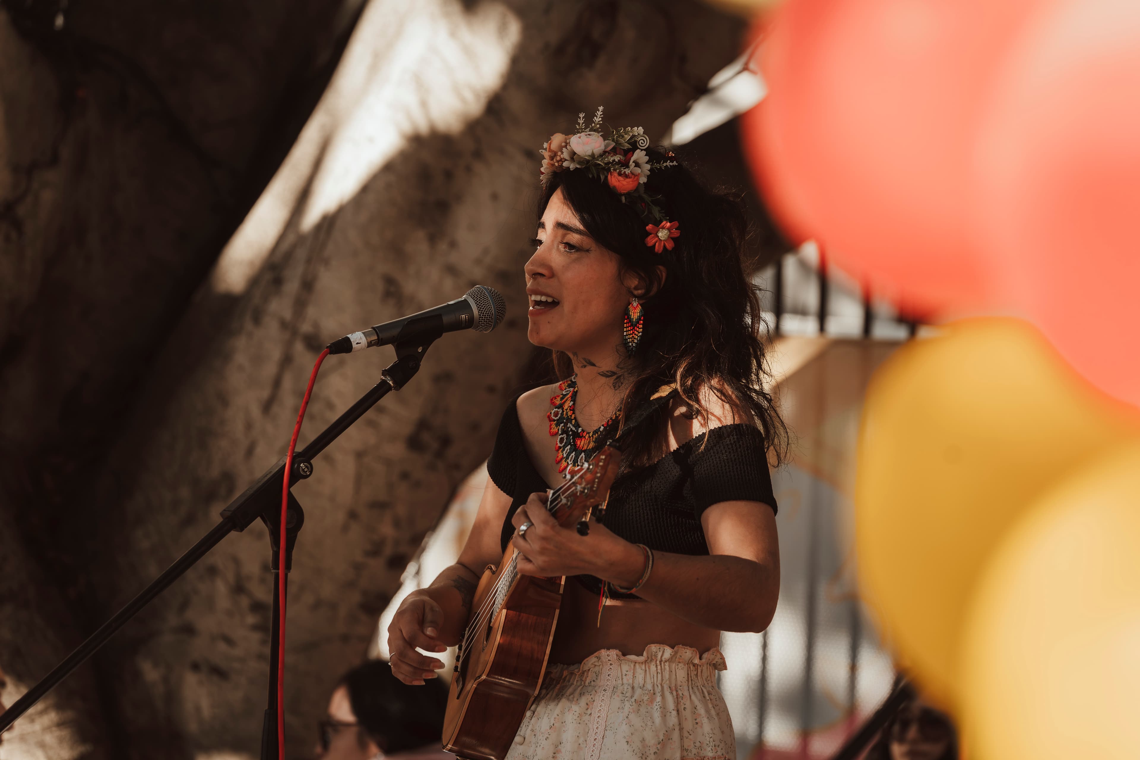 Woman wearing a flower crown sings and plays an instrument outdoors near colorful blurred balloons.
