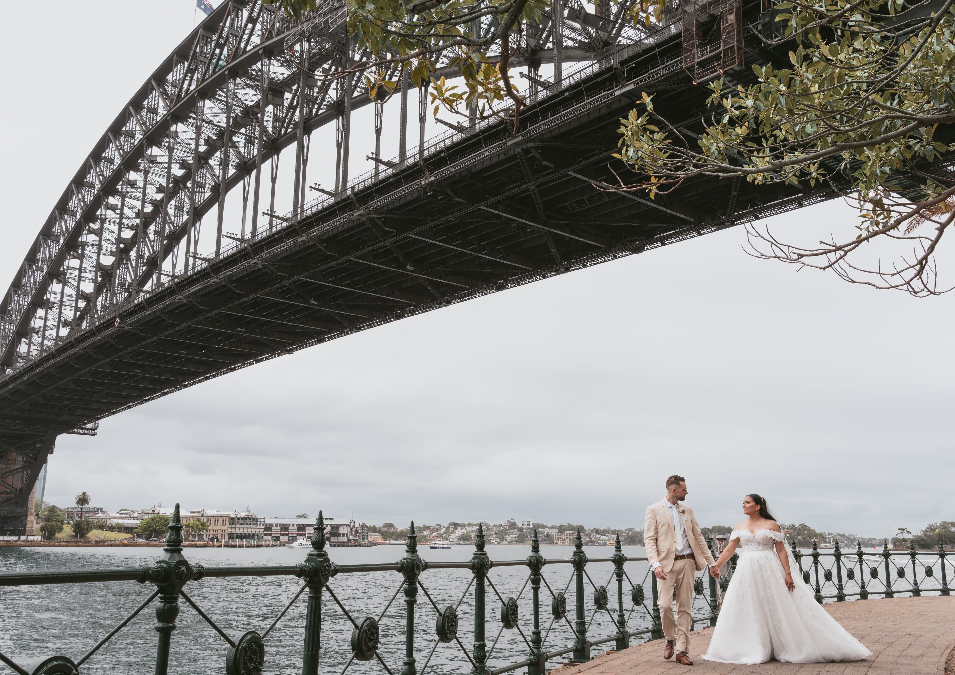 A bride and groom walk along a scenic waterfront path beneath the Sydney Harbour Bridge.