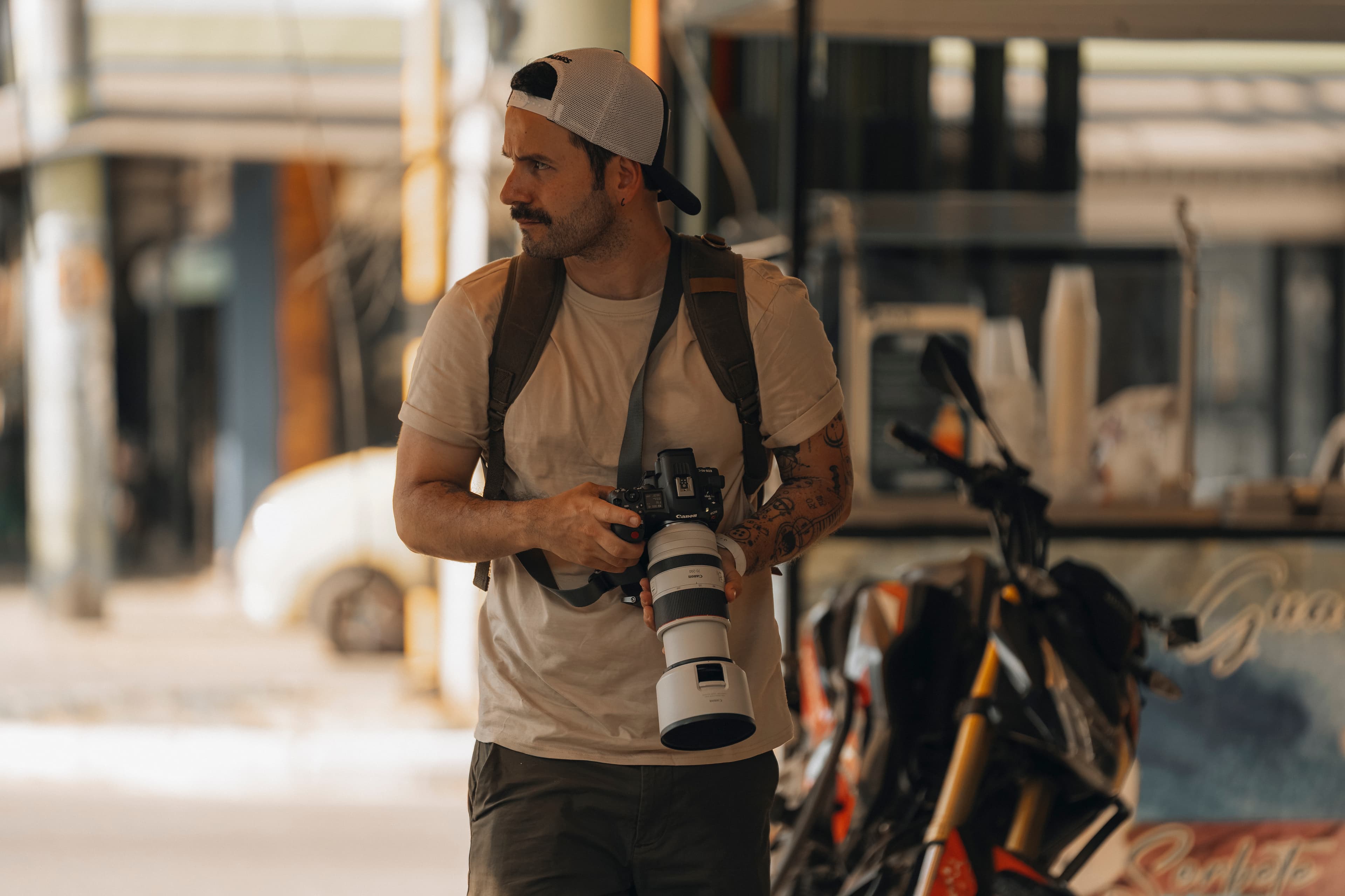 Tattooed man with a mustache holding a professional camera with a white telephoto lens.