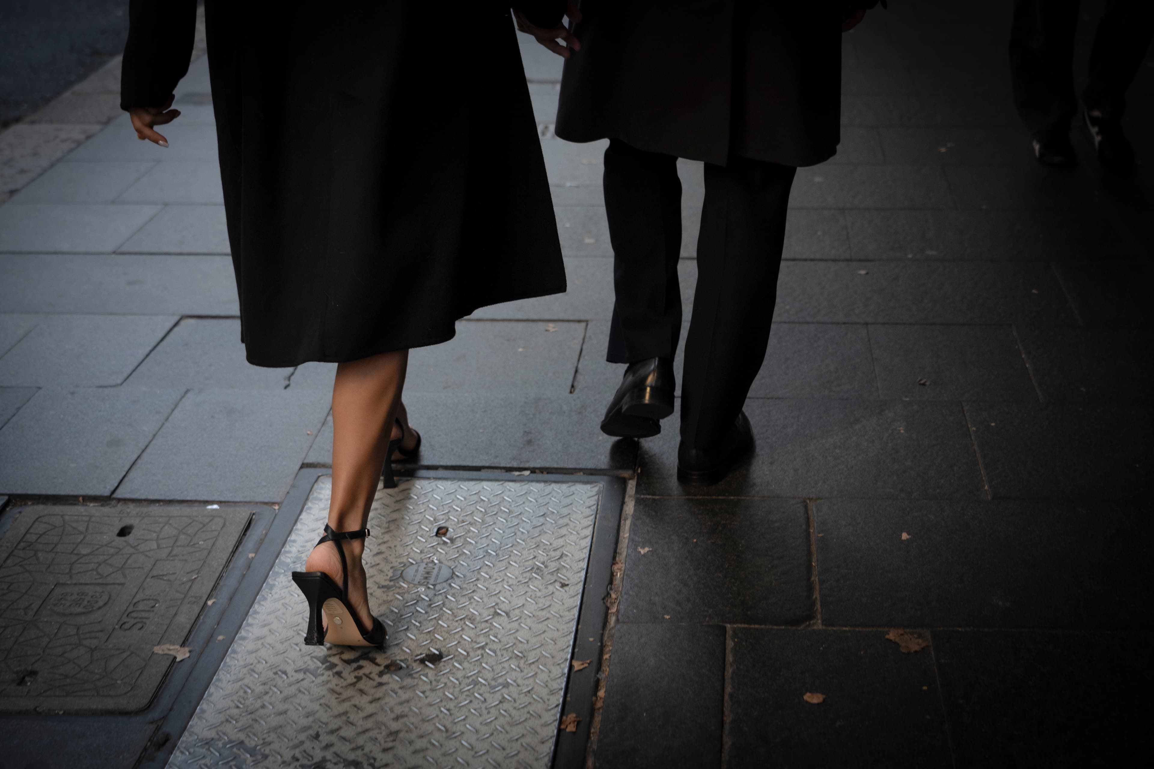 Two people in long black coats and formal shoes walk away on a stone sidewalk.