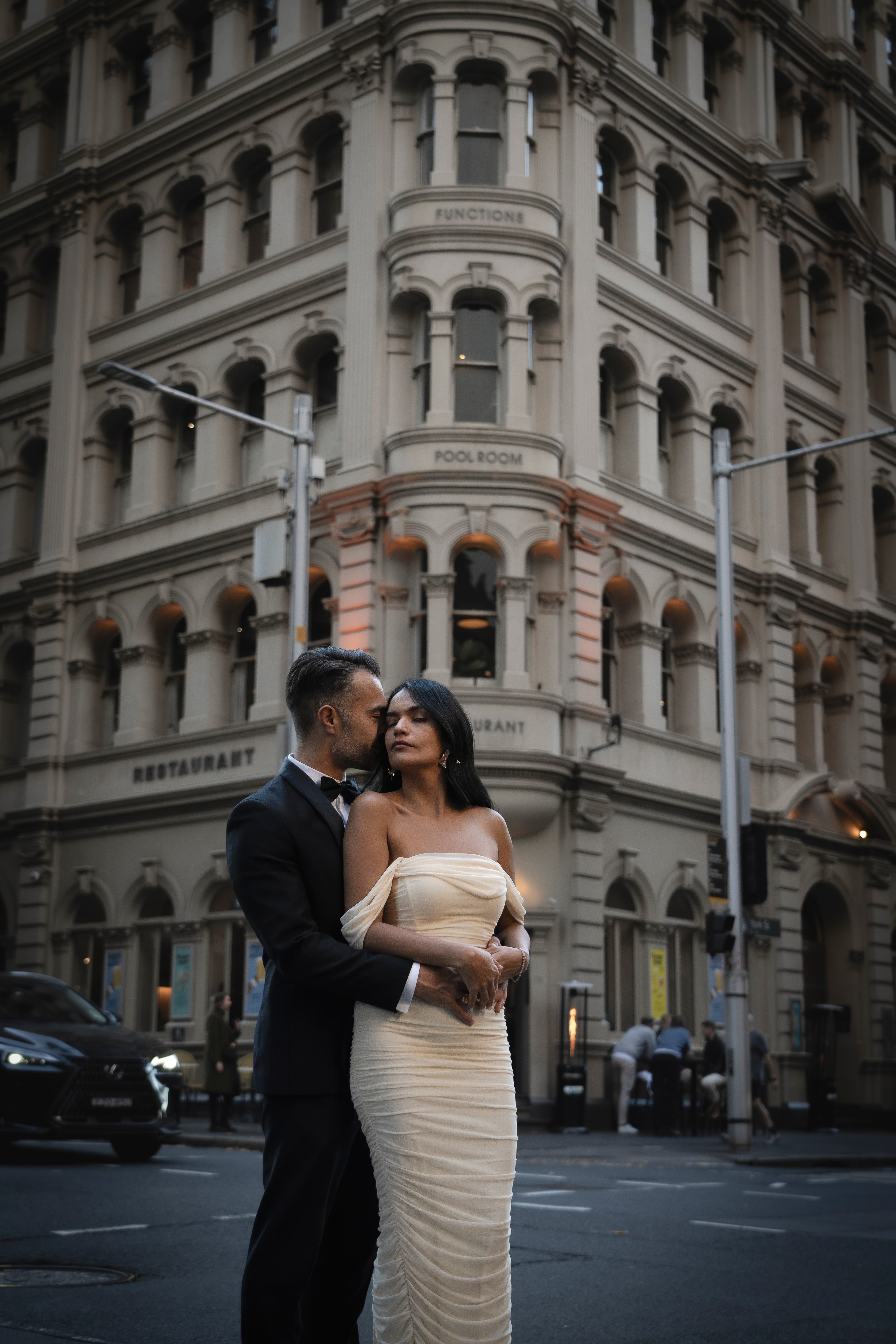 Man in tuxedo and woman in white dress embrace before a grand, ornate city building.