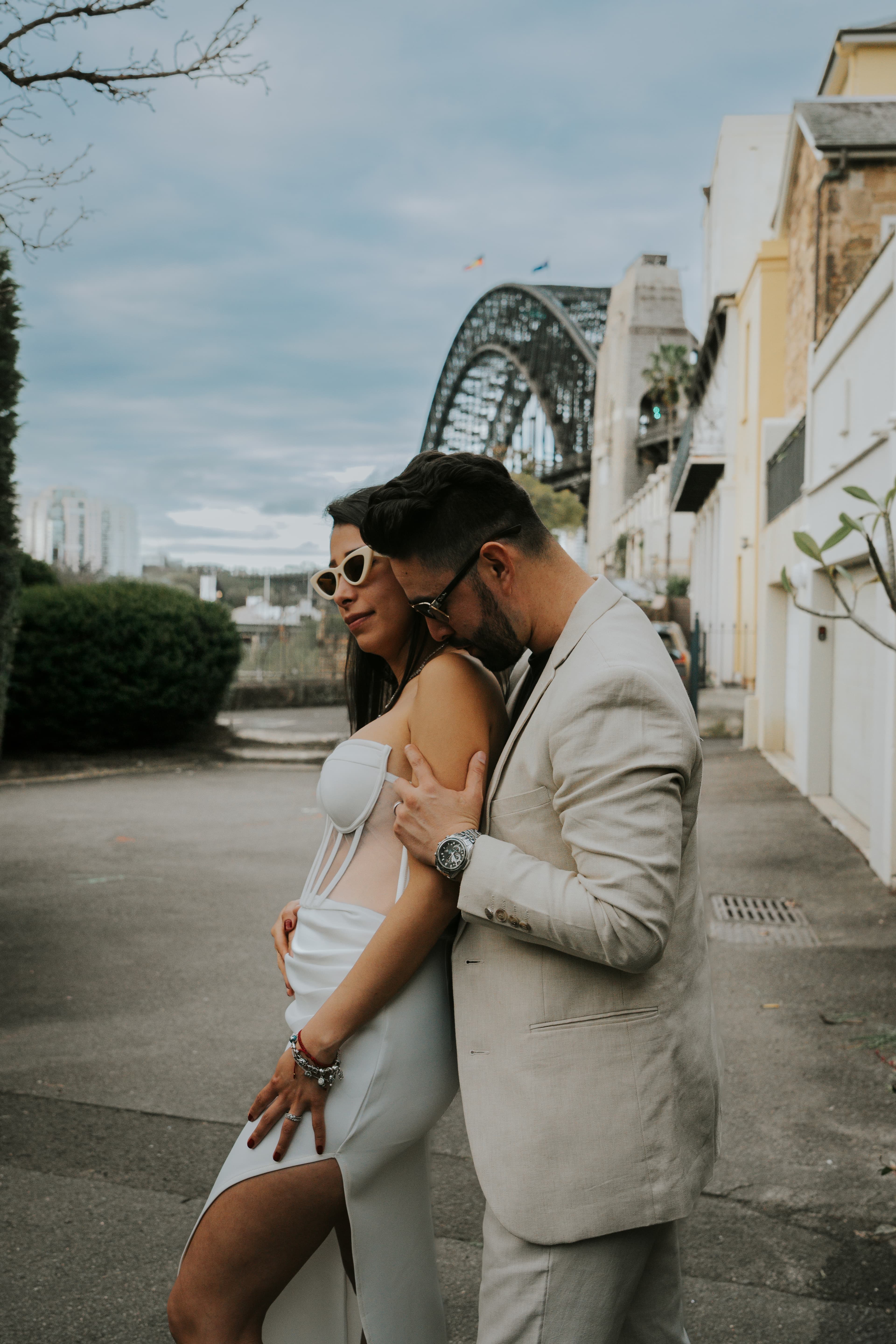 A stylish couple embraces on a street with the Sydney Harbour Bridge in the background.
