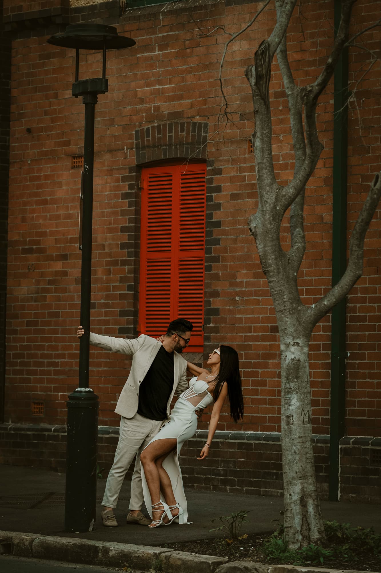 Man in light suit dips woman in white dress against brick wall with red shutters.