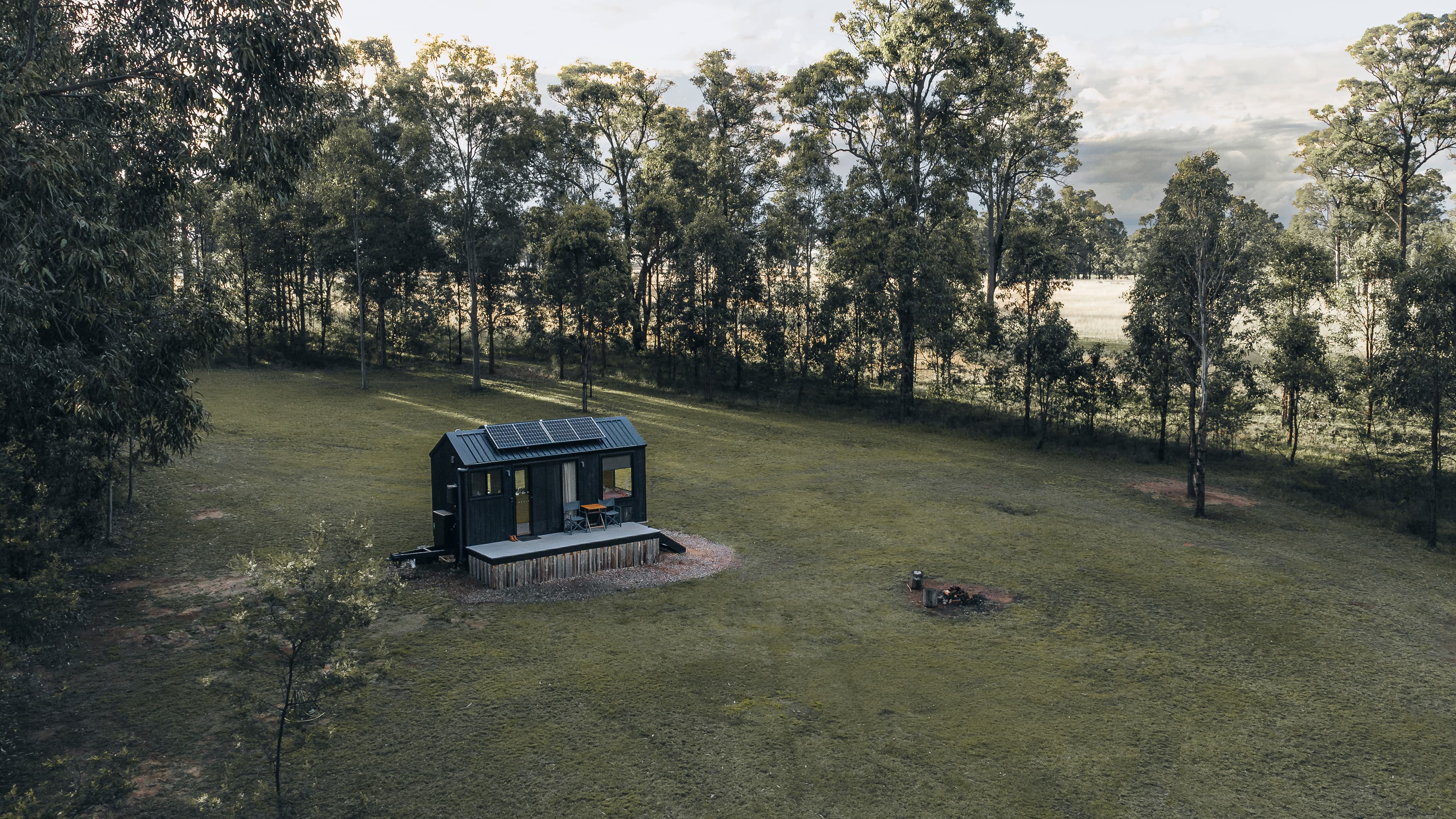 Black tiny house with solar panels in a large grassy field surrounded by tall trees.