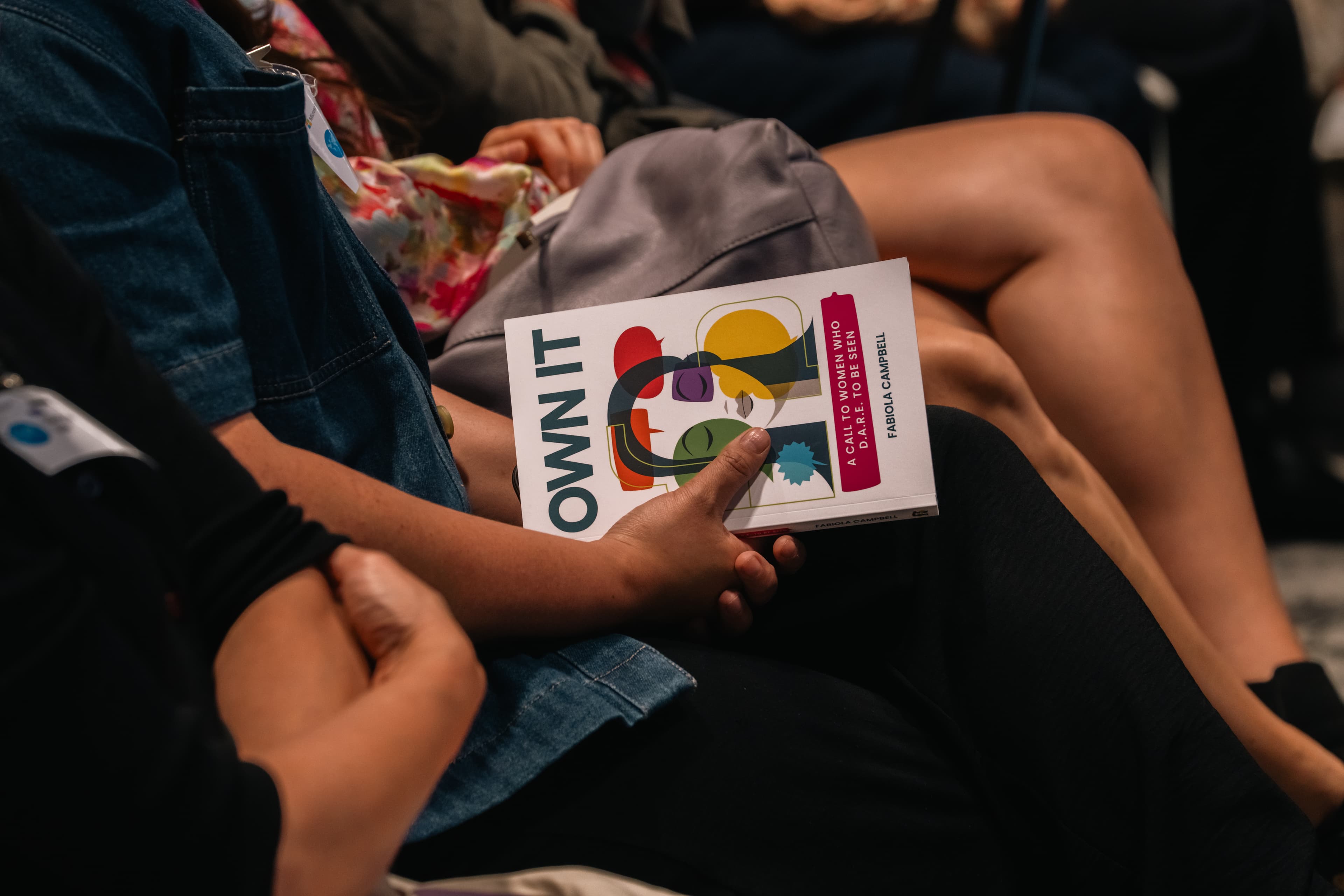 A person sitting in an audience holds the book 'OWN IT' by Fabiola Campbell.