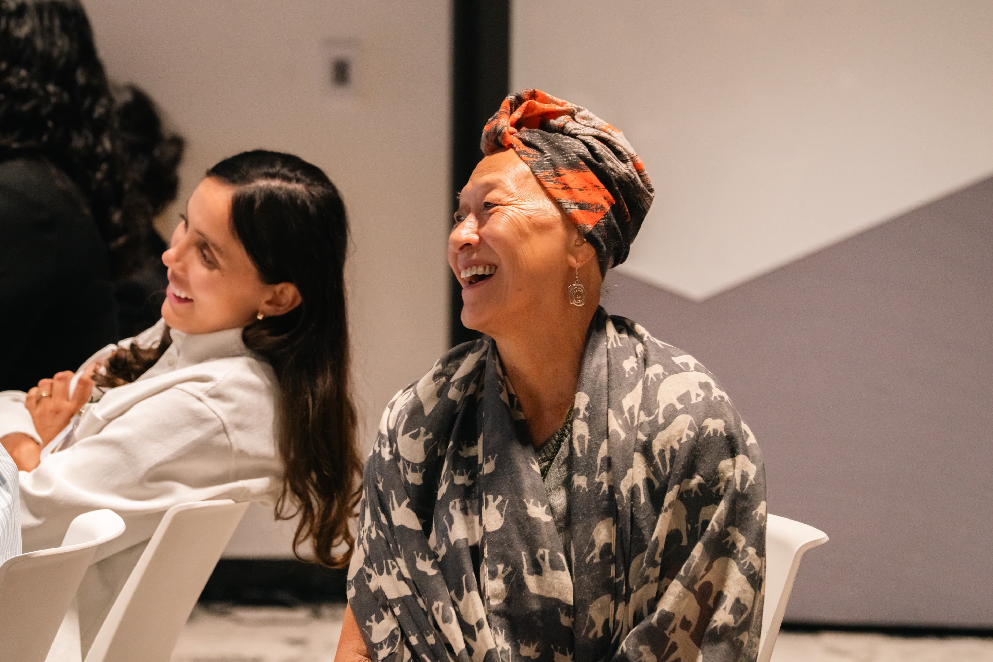 An older woman laughs heartily, wearing a vibrant headwrap and a grey elephant-patterned shawl.