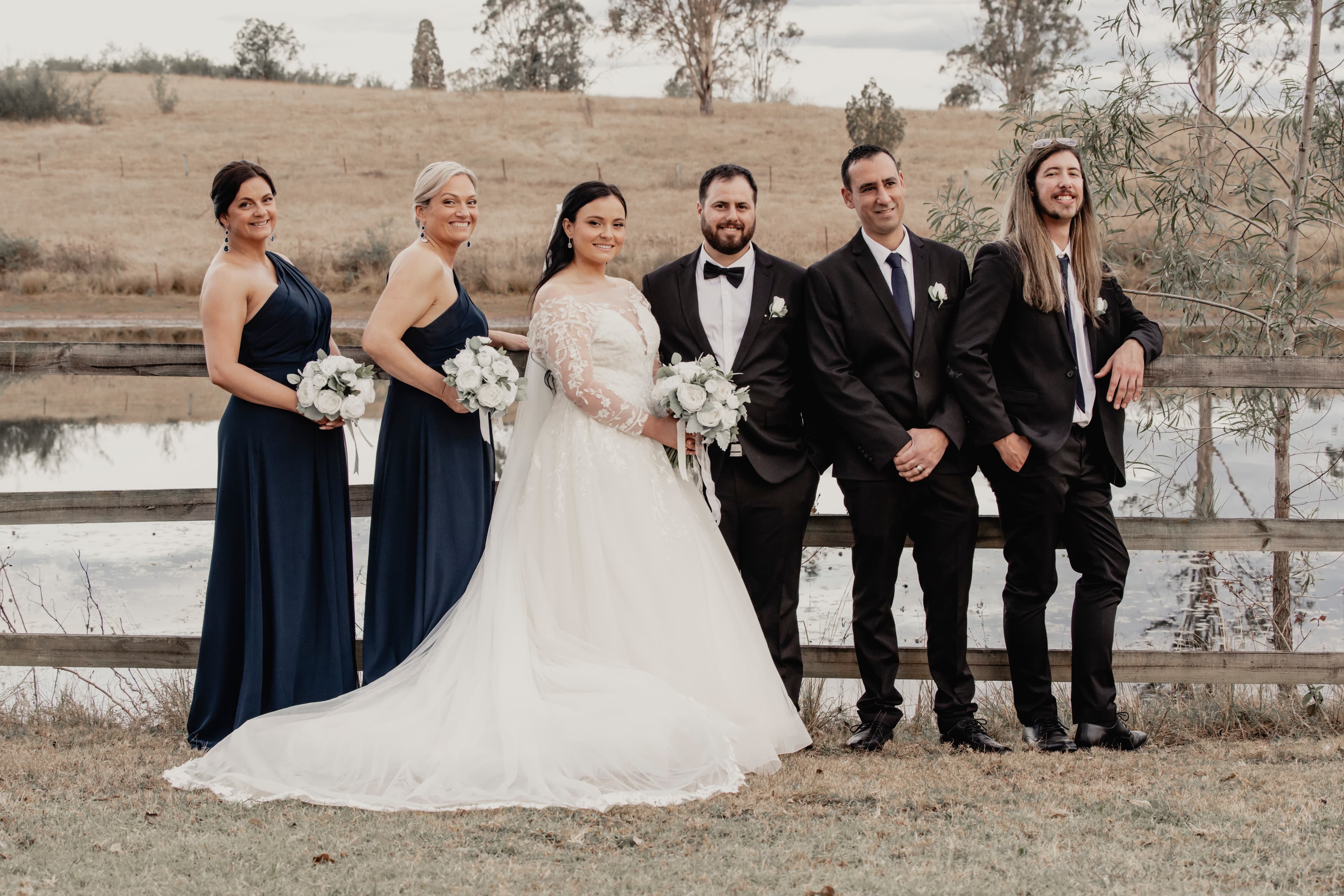 Bride, groom, and wedding party posing outdoors by a wooden fence and a scenic pond.