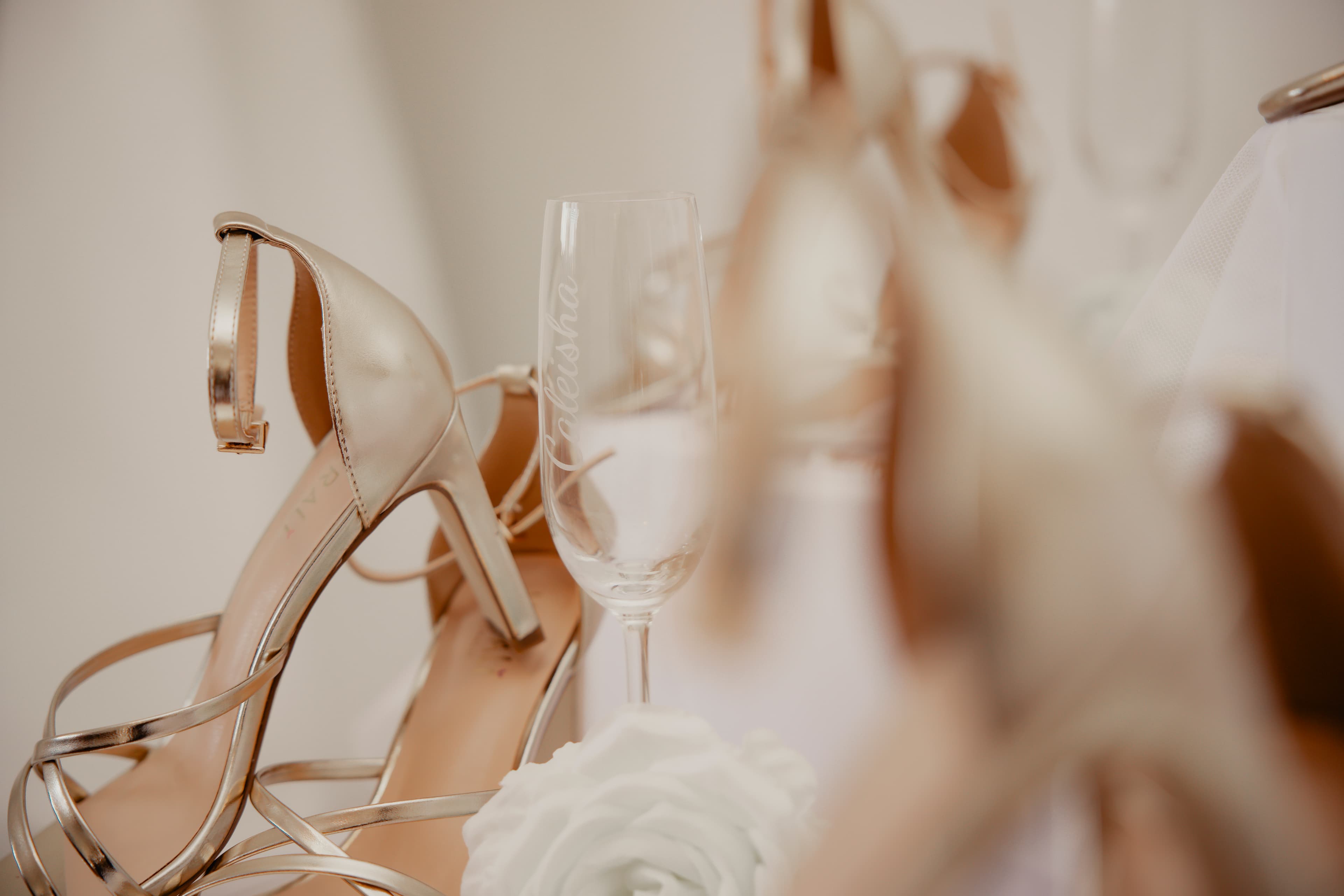 Gold strappy heels, a personalized champagne flute, and a white rose in soft focus.