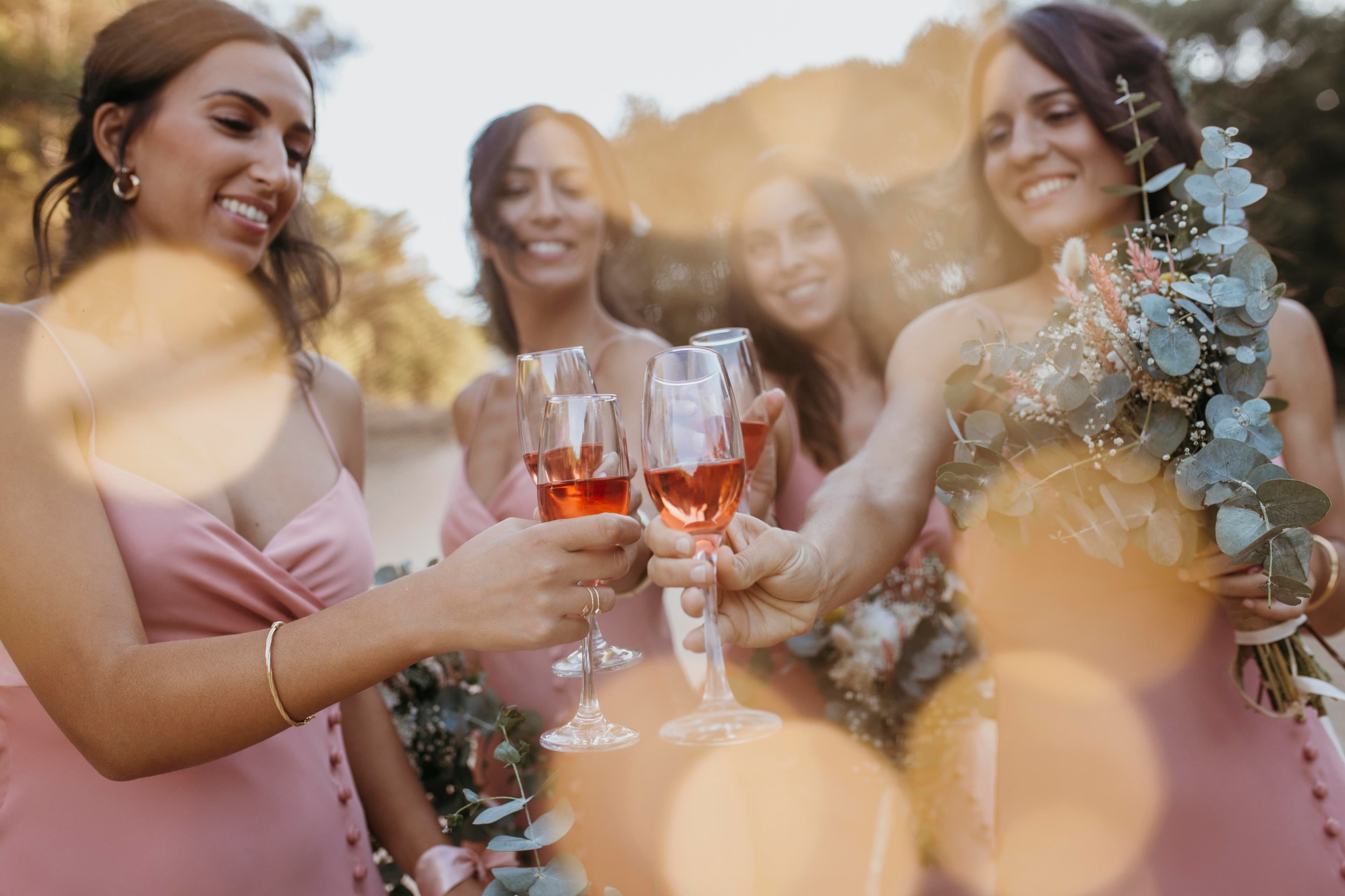 Bridesmaids in pink dresses toast with rosé wine glasses during a golden hour outdoor wedding.