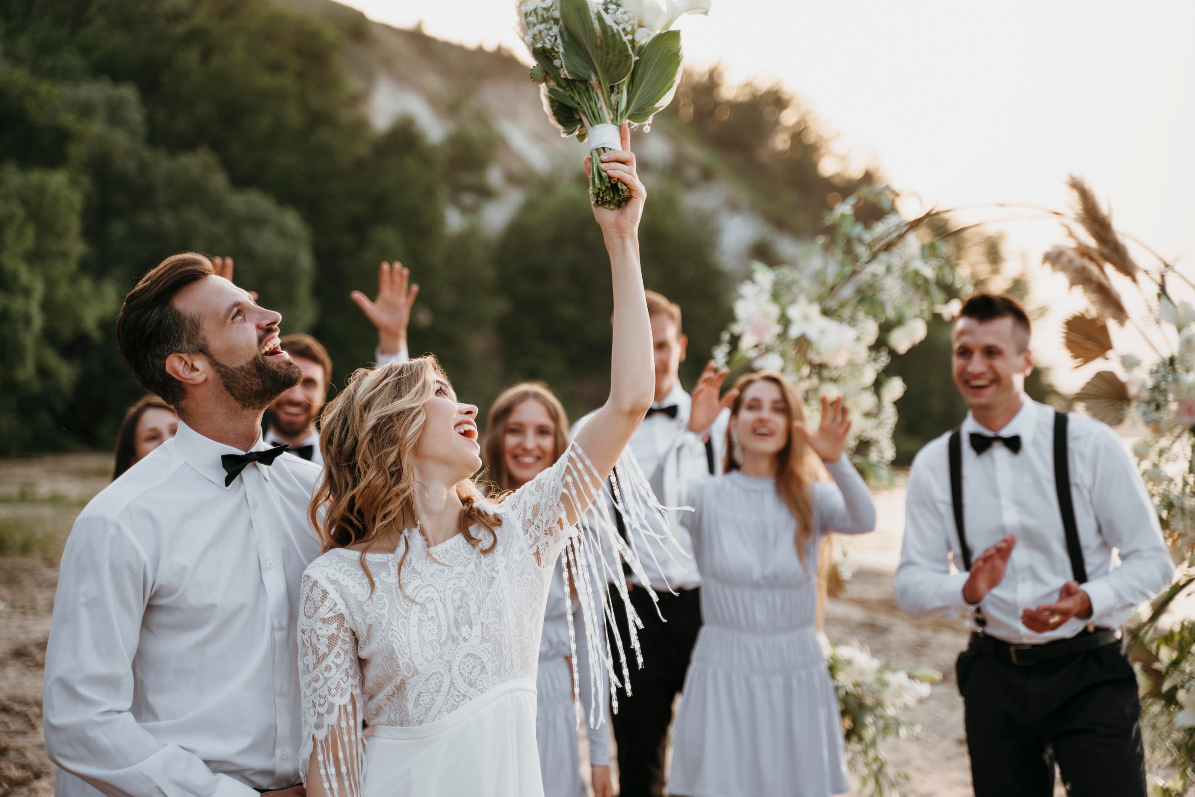 Smiling bride raises her bouquet high as the groom and wedding party celebrate outdoors.